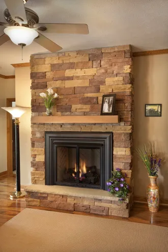 Floor-to-ceiling fireplace with multicolored stacked stone, a light wood mantel, a black metal insert with a fire, and a ceiling fan in a rustic living space.